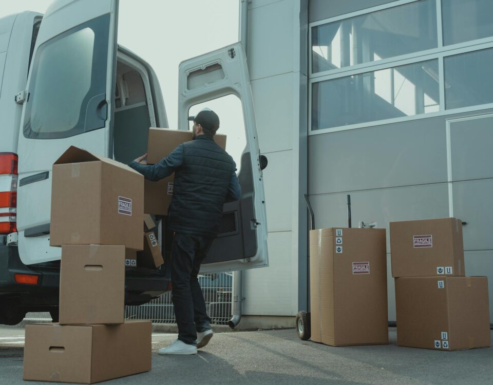 A delivery man unloading cardboard boxes from a van at a warehouse during the day.